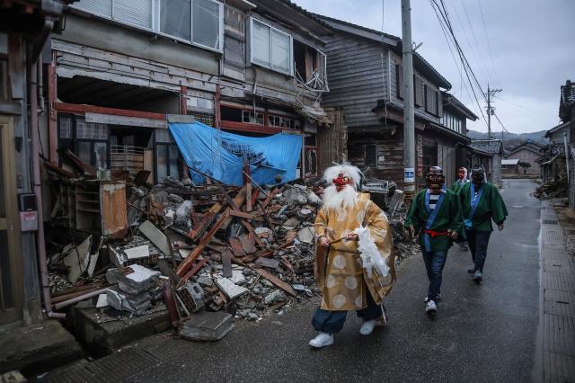 This photo taken on January 2, 2025 shows men in masks and costumes walking past homes - still badly damaged a year after the 2024 New Year's Day earthquake, as they go from house to house for the "Noto no amamehagi" tradition to drive away evil spirits, in the city of Wajima, Ishikawa prefecture. The quake on New Year's Day 2024 was Japan's deadliest in over a decade, claiming nearly 470 lives. (Photo by JIJI Press / AFP) / Japan OUT