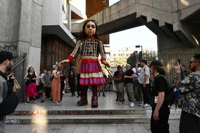 People look at Little Amal, a 12-foot-tall puppet representing a ten-year-old Syrian refugee, during a performance at the Gabriel Mistral Cultural Center as part of the opening of the 32nd version of the International Festival Teatro a Mil in Santiago on January 3, 2025. Santiago a Mil International Theatre Festival is an annual performing arts festival that takes place in January in Santiago, Chile. It is the largest arts festival in Chile and includes theatre, dance, and music events from Chile and abroad, performed in the capital city, Santiago, and around the country. (Photo by RODRIGO ARANGUA / AFP)