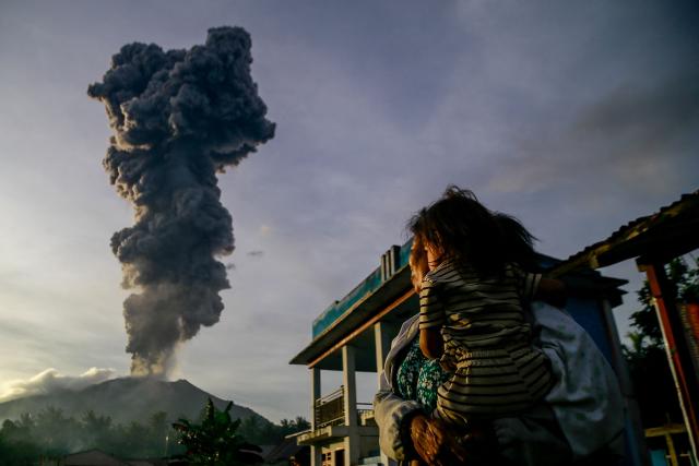 TOPSHOT - A woman and child look on at volcanic ash rising into the air during the eruption of Mount Ibu, as seen from Duono Village in West Halmahera, North Maluku province, on January 15, 2025. (Photo by AZZAM / AFP)