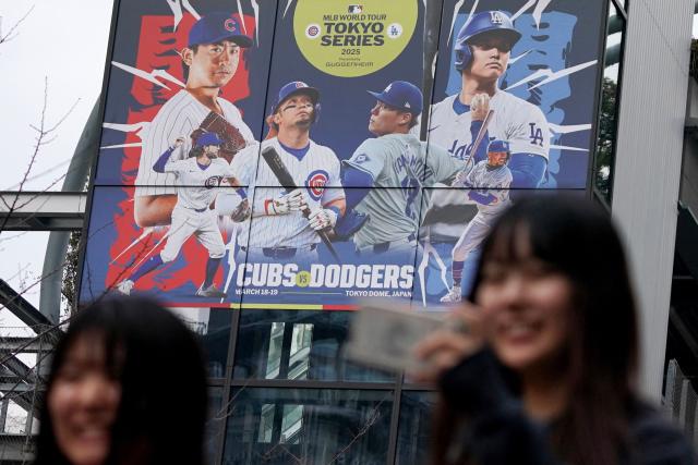 Women walk past a display showing an advertisement for the opening match of the new Major League Baseball (MLB) season between the Los Angeles Dodgers and the Chicago Cubs in the MLB Toyko series in Tokyo on March 13, 2025. Hundreds of Japanese fans waited in vain for a glimpse of Shohei Ohtani as the baseball superstar and his Los Angeles Dodgers team-mates arrived in Tokyo on March 13. (Photo by Kazuhiro NOGI / AFP)