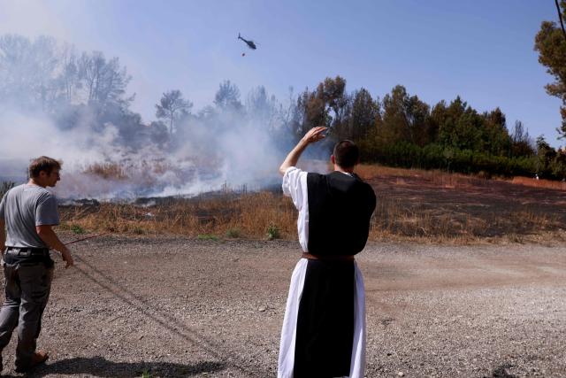 A monk stands near a man as they watch a helicopter helping to extinguish a forest fire near Latrun Monastery in central Israel, on May 1, 2025. Israeli firefighting teams battled wildfires near Jerusalem for a second day on May 1, with police reporting the reopening of several major roads that had been closed. (Photo by Menahem Kahana / AFP)