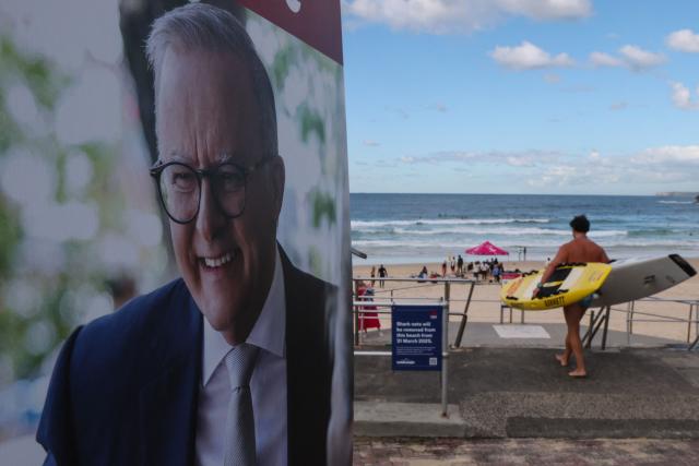 TOPSHOT - A man carries surfboards past a poster of Australia's Prime Minister Anthony Albanese for the general election in Bondi beach in Sydney on May 3, 2025. Australians began voting on May 3 in a bitterly contested general election, deciding a contest shaped by living costs, climate anxiety and US President Donald Trump's tariffs. (Photo by DAVID GRAY / AFP)