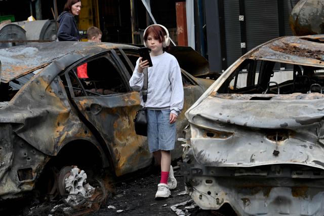 TOPSHOT - A girl listens music as she walks past burned out vehicles parked outside a shopping mall following a night-time drone attack on Kyiv, on May 4, 2025 amid the Russian invasion of Ukraine. (Photo by Sergei SUPINSKY / AFP)