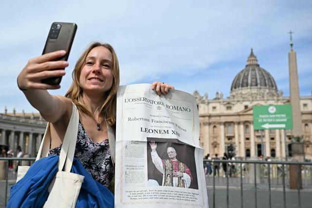 A woman poses, uses her smartphone and holds a newspaper with the picture of newly elected pope Leo XIV, with St Peter's Basilica in the background in The Vatican on May 9, 2025. (Photo by Gabriel BOUYS / AFP)