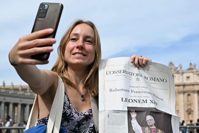A woman poses, uses her smartphone and holds a newspaper with the picture of newly elected pope Leo XIV, with St Peter's Basilica in the background in The Vatican on May 9, 2025. (Photo by Gabriel BOUYS / AFP)