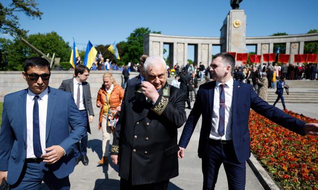 People wave Ukrainian flags (L, background) as Russia's ambassador to Germany Sergey Nechayev (C) leaves after visiting the Soviet War Memorial in Berlin's Tiergarten district during celebrations to mark the 80th anniversary of the Soviet Union's victory over Nazi Germany in the Second World War (WWII) on May 9, 2025. (Photo by Odd ANDERSEN / AFP)