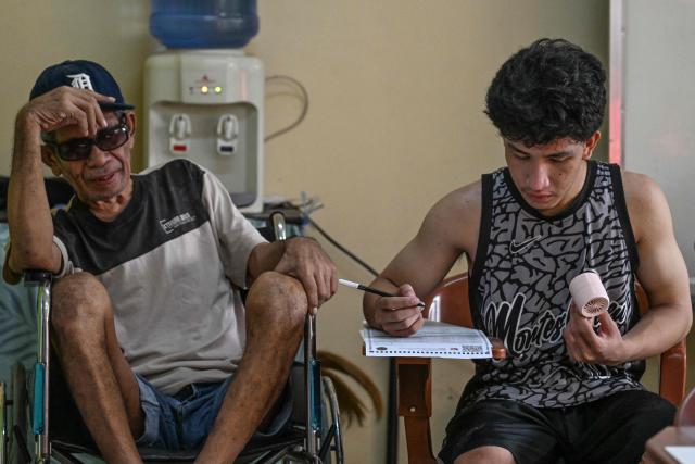 A man marks ballot papers as an older man in a wheelchair (L) looks on at a polling station during mid-term elections in Manila on May 12, 2025. Millions of Filipinos headed to the polls May 12 in a mid-term election widely seen as a referendum on the explosive feud between President Ferdinand Marcos and impeached Vice President Sara Duterte. (Photo by Jam STA ROSA / AFP)