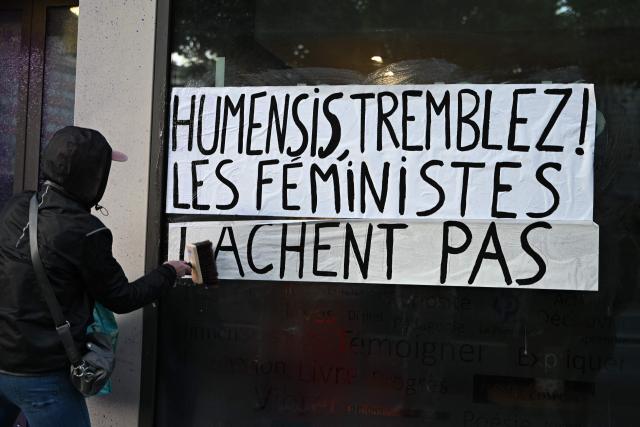 An activist from the feminist collective "NousToutes" sticks a banner reading "Humensis tremble. Feminists don't let it go" on the facade of the publishing house Humasis, to protest against the publication of  Nicolas Bedos' latest book “La soif de honte” in Paris, on May 23, 2025. French actor-writer-director Nicolas Bedos was sentenced at first instance on October 2024 to one year in prison with six months suspended for sexually assaulting two women. (Photo by Leo VIGNAL / AFP)
