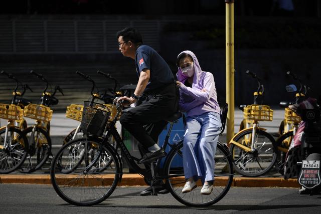A man rides a bicycle with a woman wearing sun protective clothing sitting on the back seat along a street on a hot day in Beijing on June 6, 2025. (Photo by WANG Zhao / AFP)