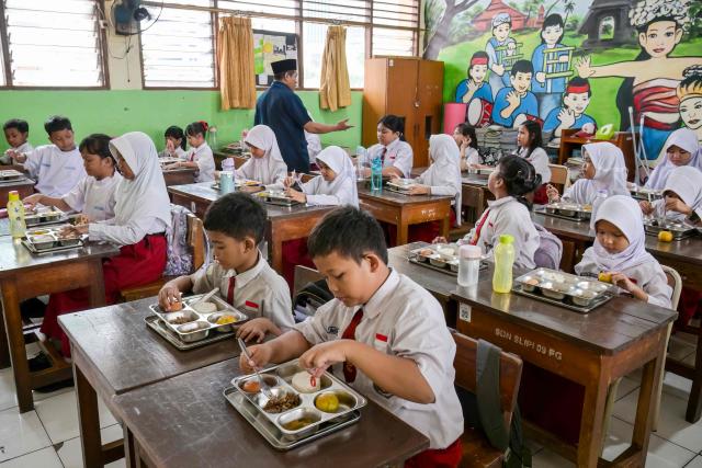 This picture taken on June 23, 2025 shows elementary schoolchildren eating food prepared by the government's free meal program at a classroom in Jakarta. Indonesian President Prabowo Subianto touted the populist scheme as a solution to the high rates of stunted growth among children, as he carved his way to a landslide election victory last year. But its rollout since January has stumbled from crisis to crisis, including accusations of nepotism, funding delays, protests and a spate of food poisonings. (Photo by BAY ISMOYO / AFP) / To go with AFP story Indonesia-politics-food-economy, FOCUS by Dessy SAGITA