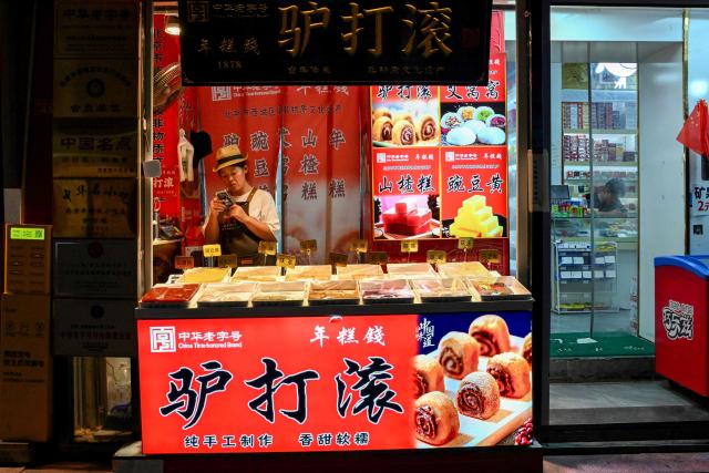 A vendor checks her cellphone while waiting for customers, in the Qianmen promenade in Beijing on July 5, 2025. (Photo by Adek BERRY / AFP)