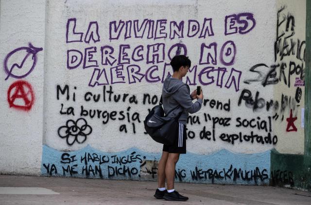 A man uses his mobile phone to record a wall with graffiti a day after a march against gentrification and the increasing presence of US citizens in the La Condesa neighbourhood of Mexico City at Mexico Park on July 5, 2025. Shouting "gringos out!" and waving banners and vandalizing various businesses, hundreds of people demonstrated this Friday in Mexico City against the gentrification of various neighborhoods due to the arrival of foreigners. (Photo by Yuri CORTEZ / AFP)