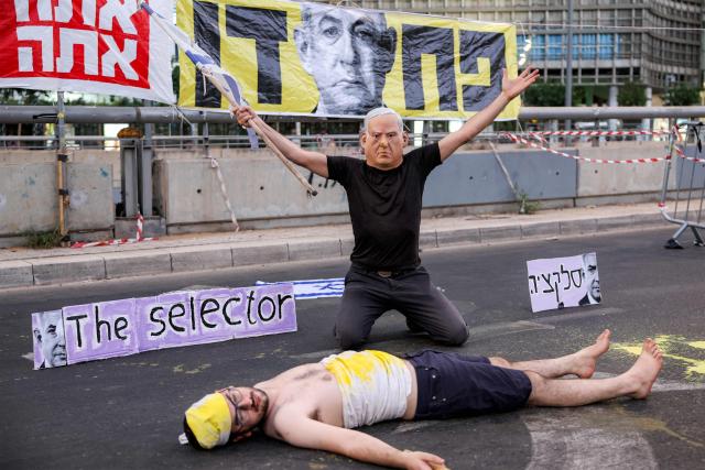 A demonstrator wearing a mask depicting Israeli Prime Minister Benjamin Netanyahu with an elongated nose, evoking the literary character Pinocchio, poses above another lying on the ground while depicting an Israeli hostage during an anti-government protest calling for action to secure the release of Israeli hostages held captive in the Gaza Strip by Palestinian militants since the 2023 October 7 attacks, outside the Israeli Defence Ministry headquarters in Tel Aviv on July 5, 2025. (Photo by Jack GUEZ / AFP)
