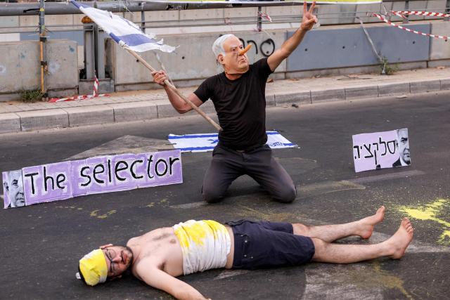 A demonstrator wearing a mask depicting Israeli Prime Minister Benjamin Netanyahu with an elongated nose, evoking the literary character Pinocchio, poses above another lying on the ground while depicting an Israeli hostage during an anti-government protest calling for action to secure the release of Israeli hostages held captive in the Gaza Strip by Palestinian militants since the 2023 October 7 attacks, outside the Israeli Defence Ministry headquarters in Tel Aviv on July 5, 2025. (Photo by Jack GUEZ / AFP)