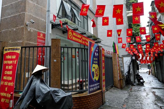 (FILES) A woman wearing a raincoat walks with her bicycle past a former local authorities' building which closed due to Vietnam's new administrative structure of 34 provinces and cities in Hanoi on July 1, 2025. Vietnam is in the midst of a dramatic remaking of its state apparatus, with 100,000 positions slated to be scrapped as Hanoi seeks to streamline bureaucracy and boost the economy. (Photo by Nhac NGUYEN / AFP)