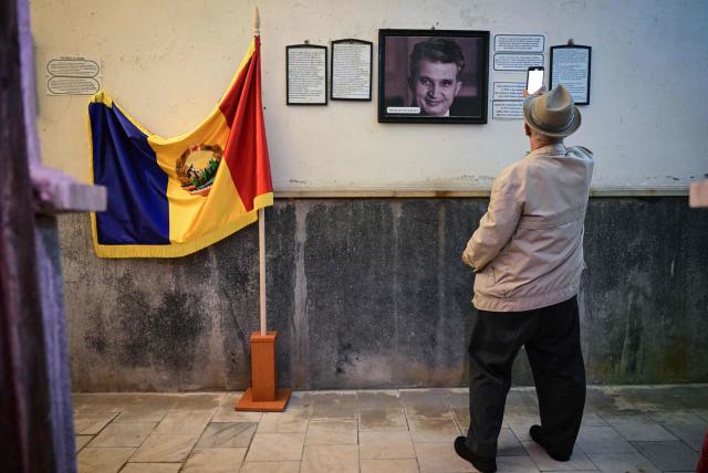 A man use his phone to enlarge the explanatory texts next to a portrait of late communist dictator Nicolae Ceausescu and a flag of socialist Romania displayed for visitors at "Apollo Baths" - a private museum in Baile Herculane, south-western Romania on July 29, 2025. A recent survey shows a worring nostalgic trend such as 55.8% of Romanians believe that the communist regime was rather a good thing for Romania, 48.4% think that life was better before 1989 compared to today, 77.2% believe that Romania was a richer country before 1989, 75.1% believe movies produced in Romania before 1989 were better than today.  President Nicusor Dan's reacts with a tweet about the survey: "I am deeply concerned about the results of the study presented today". "In my opinion, the main reason why more and more Romanians are idealizing communism is the deep disappointment with the political class of the last three decades." (Photo by Daniel MIHAILESCU / AFP)