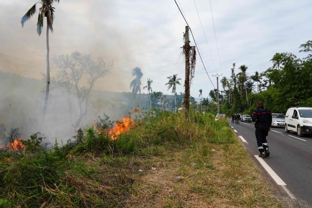 A firefighter walks along a road lined with burning vegetation during a wildfire in the village of Ironi Be, in the township of Dembeni, on the French Indian Ocean territory of Mayotte on August 12, 2025. Since Cyclone Chido hit Mayotte on December 14, 2024, fires have been fueled by fallen vegetation. (Photo by Marine GACHET / AFP)
