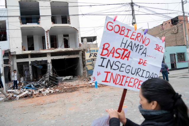 A woman holds a sign against the government as she protests in front of the remains of a house that suffered a dynamite attack caused by clashes between criminal organizations in the city of Trujillo, northern Peru on August 15, 2025. At least three people were injured and a hundred homes were affected by a dynamite attack in the Peruvian city of Trujillo, which took place during clashes between criminal organisations, authorities reported on Friday. (Photo by AFP)