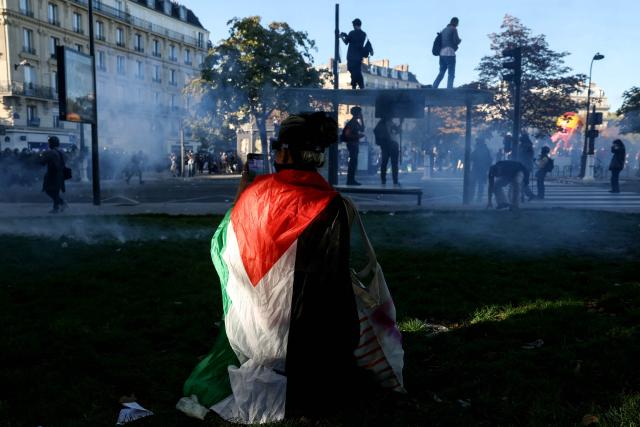 A protestor wraped in a Palestinian flag takes a photograph with her mobile phone amid the smoke in Paris on September 18, 2025, during a day of nationwide strikes and protests called by unions over France's national budget. France is bracing for a day of nationwide protests on September 18, 2025, with a source close to the authorities saying some 800,000 people are expected to take to the streets. In a rare show of unity, trade unions have urged French people to strike in protest at the authorities' "horror show" draft budget designed to reduce France's ballooning debt. (Photo by Alain JOCARD / AFP)