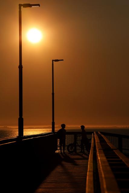 This picture taken on September 22, 2025, shows pedestrians walking on a bridge as the sun sets over Leighton Beach in Perth. (Photo by Antony DICKSON / AFP)
