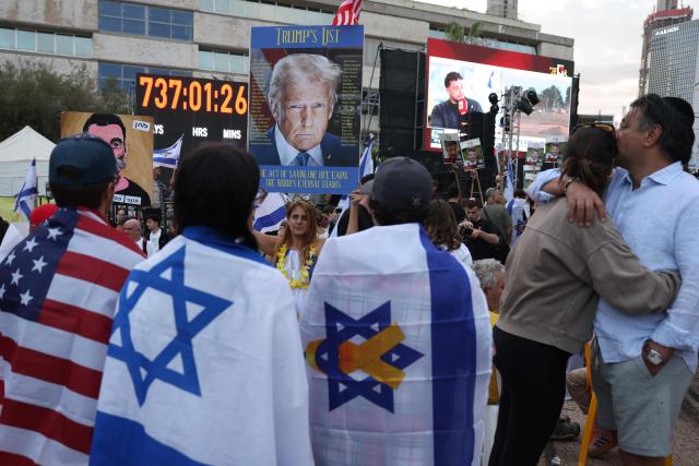 People wrapped in US and Israeli flags stand around a portrait of US President Donald Trump as they wait for the release of Israeli hostages from Gaza at Hostage Square in Tel Aviv early on October 13, 2025. Hamas was set to release all surviving hostages on on October 13 in exchange for Palestinian prisoners held by Israel, as US President Donald Trump headed to the region for a peace summit having declared the war "over". Trump's lightning visit to Israel and Egypt aims to celebrate his role in brokering last week's ceasefire and hostage release deal -- but comes at a precarious time as Israel and Hamas negotiate what comes next. (Photo by AHMAD GHARABLI / AFP)