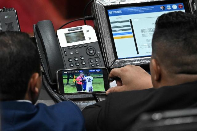 Two deputies watch the football match between the national teams of El Salvador and Guatemala on a mobile phone during a session where they might review and approve an anti-gang law at the National Congress in Guatemala City on October 14, 2025. Twenty members of a gang designated a "foreign terrorist organization" by the United States have escaped from detention in Guatemala, a prison chief said Sunday. (Photo by JOHAN ORDONEZ / AFP)