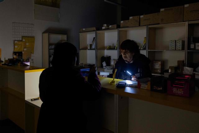 A national postal service of Ukraine worker uses a flashlight to serve a customer during a power outage in Kyiv, on October 16, 2025, amid the Russian invasion of Ukraine. (Photo by Tetiana DZHAFAROVA / AFP)