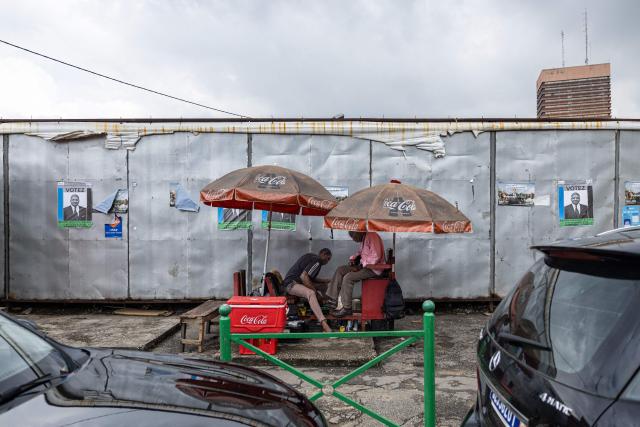 Posters of Democratic Congress (CODE) presidential candidate Jean-Louis Billon are displayed on a wall as a shoeshiner polishes a client’s shoes in Plateau near Abidjan on October 17, 2025. (Photo by EMMANUEL CROSET / AFP)