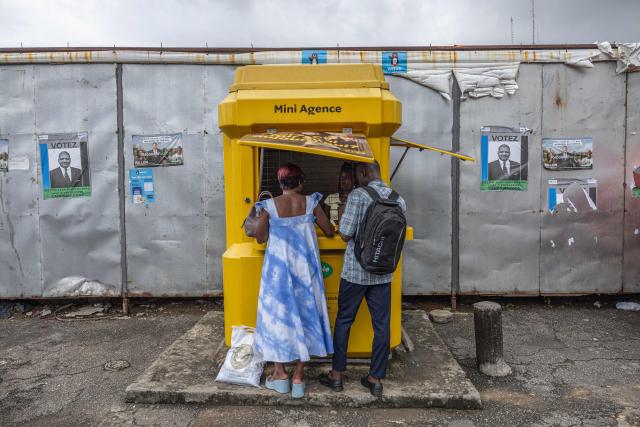 Posters of Democratic Congress (CODE) presidential candidate Jean-Louis Billon are displayed on a wall as clients make a purchase at a street kiosk in Plateau near Abidjan on October 17, 2025. (Photo by EMMANUEL CROSET / AFP)