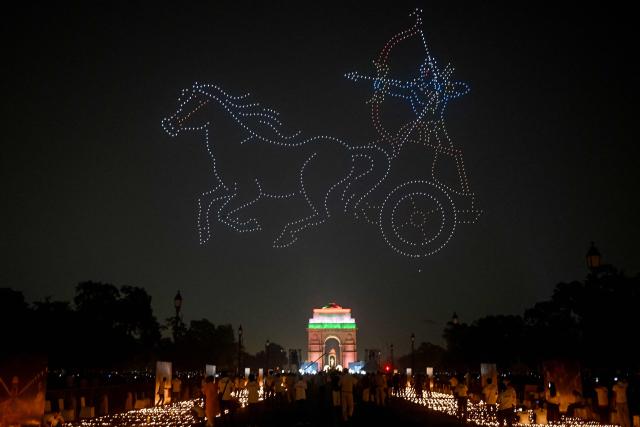People light diyas, or traditional earthen oil lamps, as illuminated drones depicting stories from the Hindu mythological epic Ramayana fly over the India Gate memorial as part of the Diwali celebrations, the Hindu festival of lights, at Kartavya Path in New Delhi on October 18, 2025. (Photo by Arun SANKAR / AFP)