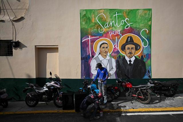 Two men repair a motorcycle next to a mural depicting Venezuelan Sister Maria (L) and Venezuelan doctor Jose Gregorio Hernandez on a street in the La Pastora neighborhood of Caracas on October 18, 2025. Pope Leo XIV is set to canonize Venezuela’s first saints, Dr. Jose Gregorio Hernandez and Sister Maria del Carmen Rendiles, during a ceremony in St. Peter's Square on October 19, 2025. (Photo by Federico PARRA / AFP)
