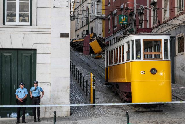 (FILES) Police officers stand behind a security cordon on the site of the Gloria funicular the day after an accident that killed 16 a day earlier in Lisbon, on September 4, 2025. An official inquiry called Monday for all Lisbon cable cars to be kept out of service for urgent modifications after a deadly crash in September that it blamed on a defective cable. The September 3 crash when the funicular came off the rails and hurtled into a building left 16 dead. The official inquiry's preliminary report said Lisbon's cable cars cannot resume operating until they can guarantee they have braking systems "capable of immobilizing the cabins in the event of a cable break." (Photo by PATRICIA DE MELO MOREIRA / AFP)