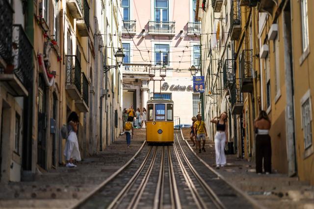 (FILES) Tourists take pictures in front of the Bica funicular, closed temporarily after the Gloria funicular accident in Lisbon on September 9, 2025. An official inquiry called Monday for all Lisbon cable cars to be kept out of service for urgent modifications after a deadly crash in September that it blamed on a defective cable. The September 3 crash when the funicular came off the rails and hurtled into a building left 16 dead. The official inquiry's preliminary report said Lisbon's cable cars cannot resume operating until they can guarantee they have braking systems "capable of immobilizing the cabins in the event of a cable break." (Photo by PATRICIA DE MELO MOREIRA / AFP)