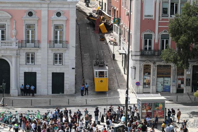(FILES) People gather to see the wreckage (up) of the Gloria funicular the day after an accident that killed 16 a day earlier in Lisbon, on September 4, 2025. An official inquiry called Monday for all Lisbon cable cars to be kept out of service for urgent modifications after a deadly crash in September that it blamed on a defective cable. The September 3 crash when the funicular came off the rails and hurtled into a building left 16 dead. The official inquiry's preliminary report said Lisbon's cable cars cannot resume operating until they can guarantee they have braking systems "capable of immobilizing the cabins in the event of a cable break." (Photo by PATRICIA DE MELO MOREIRA / AFP)