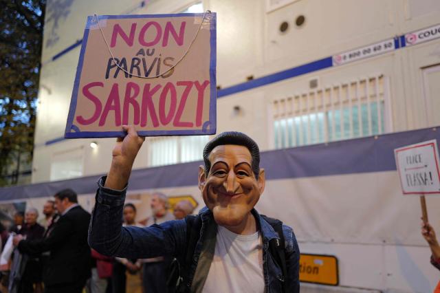 A local resident, wearing a mask of Nicolas Sarkozy, holds a placard protesting against the mayor of Nice's proposal to name the square in front of the future police station after former French President Nicolas Sarkozy, in Nice on october 21, 2025. France's fomer President Nicolas Sarkozy became the first former head of an EU state to be jailed on October 21, 2025, proclaiming his innocence as he entered a Paris prison. (Photo by Valery HACHE / AFP)