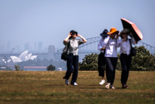 Tourists hold onto their hats and hold an umbrella as they walk in a park overlooking Sydney Harbour on a hot and windy spring day in Sydney on October 22, 2025. (Photo by DAVID GRAY / AFP)