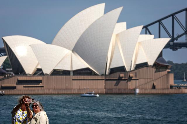 Tourists take a selfie as they stand in front of the Sydney Opera House and Harbour Bridge on a hot and windy spring day in Sydney on October 22, 2025. (Photo by DAVID GRAY / AFP)