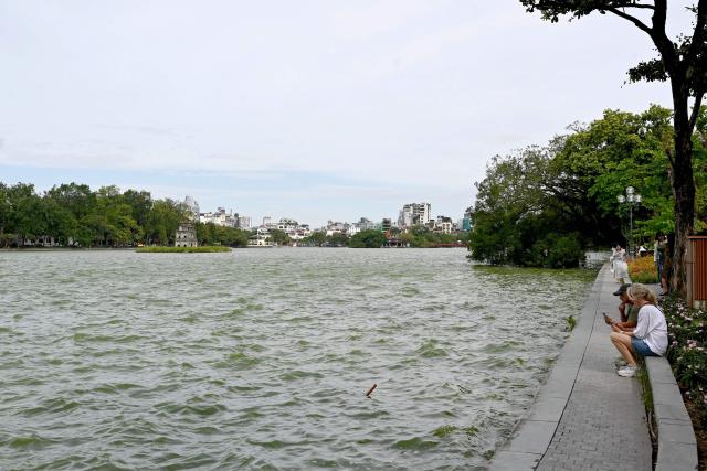 Tourists sit and use their phones on the banks of Hoan Kiem Lake in Hanoi on October 22, 2025. (Photo by Nhac NGUYEN / AFP)