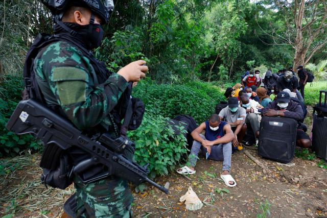 A soldier stands guard as people from various countries, who were working in the KK Park compound in Myanmar and crossed to Thailand via the Moei river, sit with their belongings in Mae Sot District, Tak Province area on October 24, 2025. More than 1,000 people have fled from Myanmar into Thailand this week, Thai authorities said on October 24, after the Myanmar military raided one of the country's largest scam centres. (Photo by Sarot Meksophawannakul / THAI NEWS PIX / AFP)