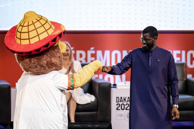 Senegal's President Bassirou Diomaye Faye (R) greets the official mascot Ayo for the Dakar 2026 Youth Olympics Games during the unveiling ceremony at the Grand Theatre in Dakar, on October 31, 2025. Senegal will be the first African country to host the Summer Youth Olympic Games in 2026. (Photo by PATRICK MEINHARDT / AFP)