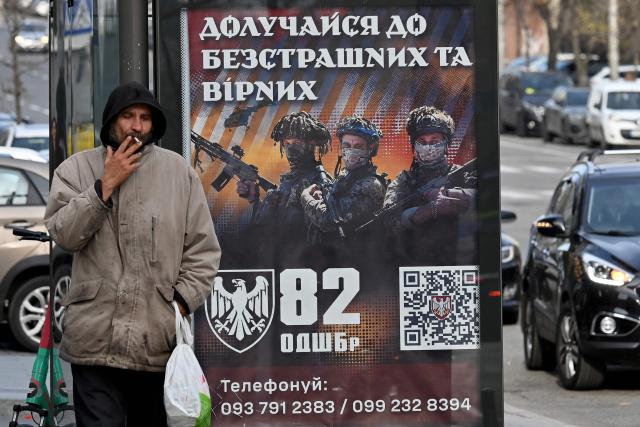 A man smokes in front of a recruiting placard  of the 82 separate assault brigade which reads "Join the fearless and faithful" at a bus stop in Kyiv on November 2, 2025, amid the Russian invasion of Ukraine. (Photo by Sergei SUPINSKY / AFP)