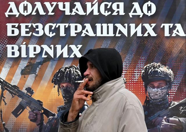 A man smokes in front of a recruiting placard  of the 82 separate assault brigade which reads "Join the fearless and faithful" at a bus stop in Kyiv on November 2, 2025, amid the Russian invasion of Ukraine. (Photo by Sergei SUPINSKY / AFP)