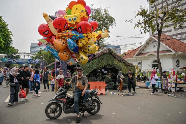 A balloon vendor waits for costumers as visitors take pictures with military vehicles during the Indonesian Marine Fair, marking the 80th anniversary of the Indonesian Marine Corps, along a road in Surabaya on November 9, 2025. (Photo by JUNI KRISWANTO / AFP)