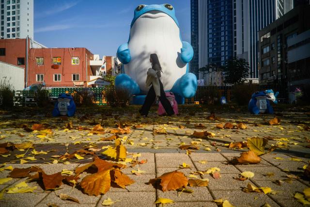 A woman walks past fallen autumn leaves on a pavement and an inflatable figure in Seoul on November 13, 2025. (Photo by ANTHONY WALLACE / AFP)