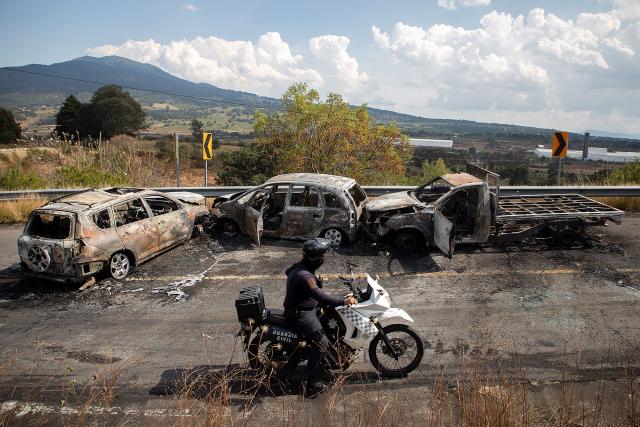 TOPSHOT - An agent of the National Guard of Mexico inspects the area where four vehicles were burned by alleged members of organized crime on a highway near Quiroga, Michoacan state, Mexico on November 17, 2025. Armed civilians blocked several roads in the western Mexican state of Michoacan on November 17, 2025, in response to an operation against a criminal leader, a week after the government of President Claudia Sheinbaum reinforced the presence of federal forces in the state. (Photo by Enrique CASTRO / AFP)