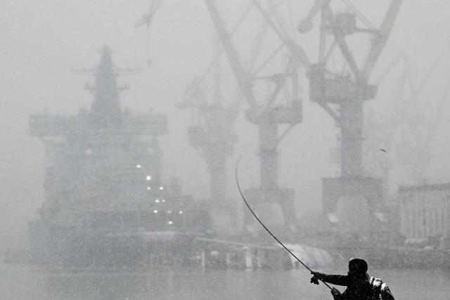 A man fishes on the Neva River embankment, with the new under construction Project 22220 Chukotka nuclear-powered icebreaker in the background, during a snowfall in Saint Petersburg on November 18, 2025. (Photo by Olga MALTSEVA / AFP)