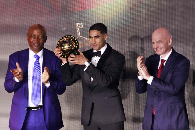 Paris Saint-Germain's Moroccan defender Achraf Hakimi (C) holds his Africa Player of the Year award next to CAF President Patrice Motsepe (L) and FIFA President Gianni Infantino (R) during the 2025 Confederation of African Football (CAF) Awards in Sale, Morocco on November 19, 2025.  (Photo by Abdel Majid BZIOUAT / AFP)