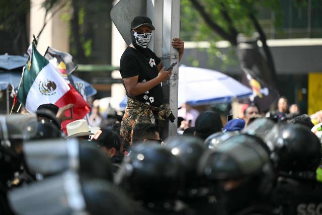 A protester wearing a skull facemask looks at riot police during a Generation Z rally against the government of Mexico's President Claudia Sheinbaum, along Reforma Avenue in Mexico City, on November 20, 2025. This is the second demonstration called by Generation Z to protest against the leftist government's security policy, after another protest on Saturday ended with more than 100 injured, most of them police officers, and 19 arrested. (Photo by Yuri CORTEZ / AFP)