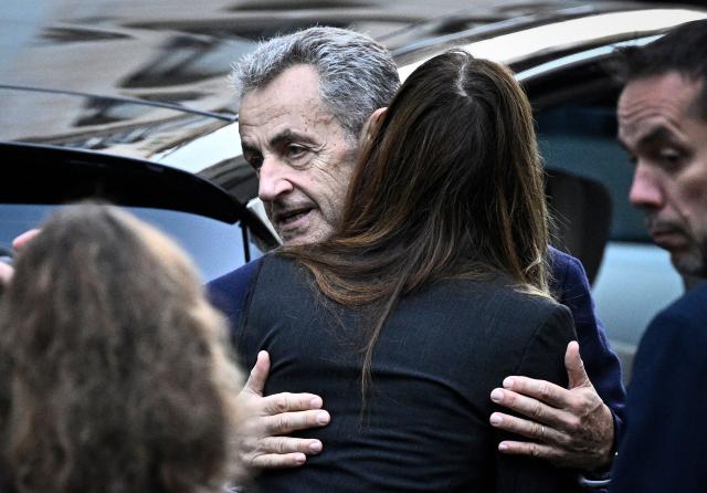 (FILES) France's former president Nicolas Sarkozy hugs his wife Carla Bruni-Sarkozy as he leaves his residence to present himself to La Sante Prison for incarceration on a five-year prison sentence after being convicted of criminal conspiracy over a plan for late Libyan dictator Moamer Kadhafi to fund his 2007 electoral campaign, in Paris, on October 21, 2025. Less than two weeks after his release from prison, Nicolas Sarkozy announced on November 21, the release on December 10 of his new book on his incarceration at Paris' Prison de la Sante. Nicolas Sarkozy was released under judicial supervision on November 10, pending an appeals trial over allegations that he sought Libyan funding. (Photo by JULIEN DE ROSA / AFP)