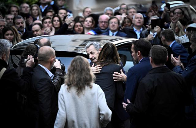 (FILES) France's former president Nicolas Sarkozy (C) hugs his wife Carla Bruni-Sarkozy as he leaves his residence to present himself to La Sante Prison for incarceration on a five-year prison sentence after being convicted of criminal conspiracy over a plan for late Libyan dictator Moamer Kadhafi to fund his 2007 electoral campaign, in Paris, on October 21, 2025. Less than two weeks after his release from prison, Nicolas Sarkozy announced on November 21, the release on December 10 of his new book entitled "Journal d'un Prisonnier" (Journal of a Prisoner) on his incarceration at Paris' Prison de la Sante. Nicolas Sarkozy was released under judicial supervision on November 10, pending an appeals trial over allegations that he sought Libyan funding. (Photo by JULIEN DE ROSA / AFP)
